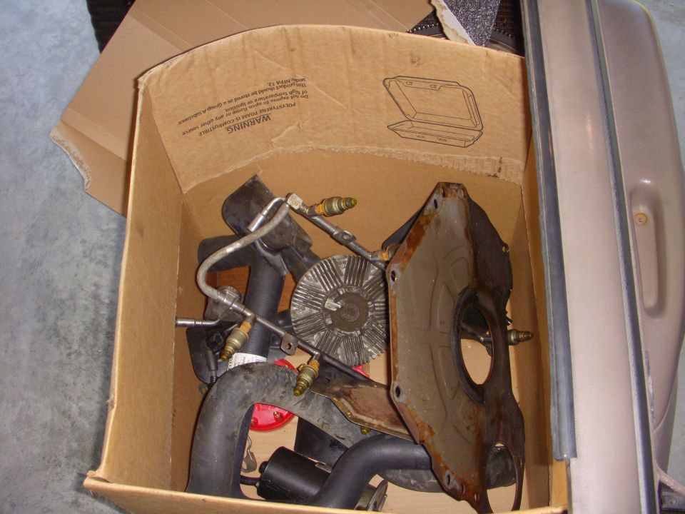 Cardboard box of used engine parts: rusty plate, fan clutch, hoses, spark plugs, and metal lines