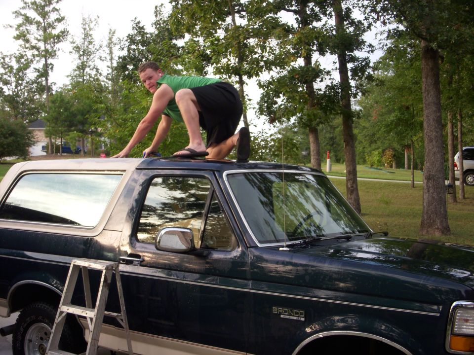 Man climbing on the roof of a dark green 1993 Ford Bronco with a ladder nearby