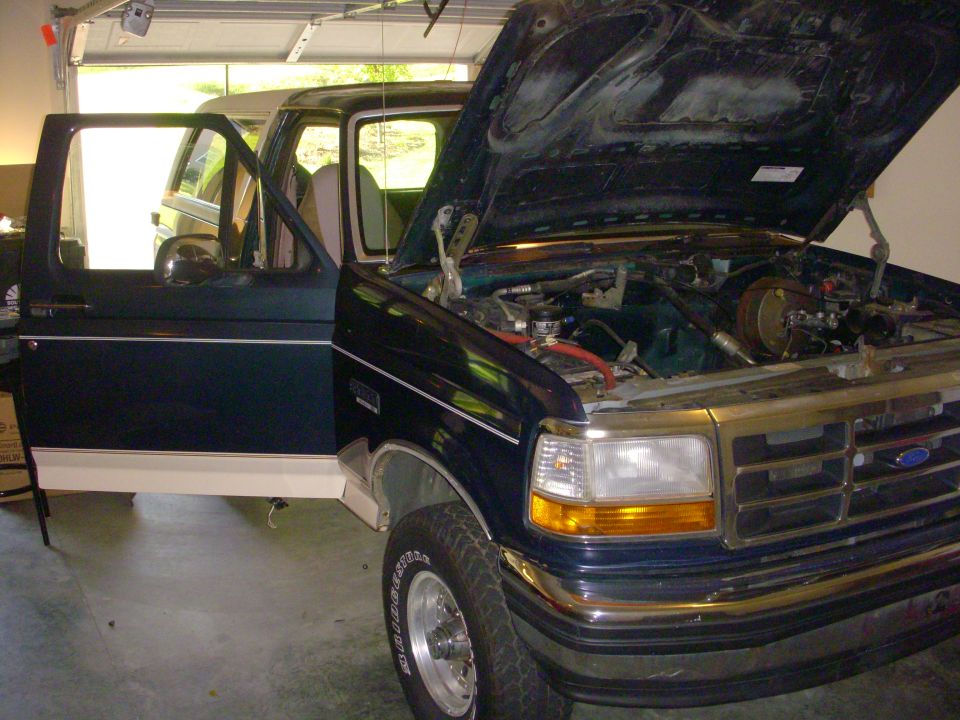 1993 Ford Bronco with driver door open and hood up, showing engine bay. Another SUV is visible in the background