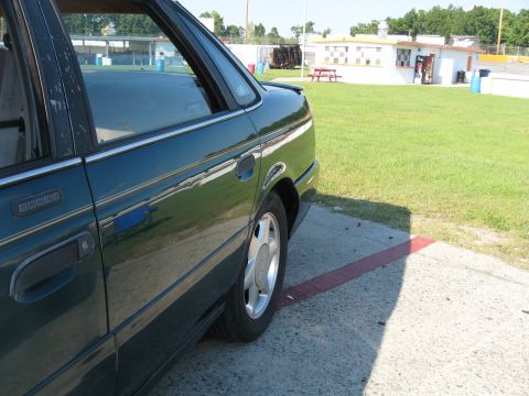 1993 Ford Taurus - The side profile of a sedan shows its rear door, window, and wheel. A building and grassy field are visible behind it.