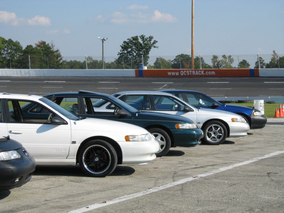 White sedan parked on asphalt, side view, black rims, sunroof, racing track in background