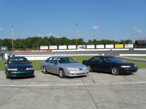 1993 Ford Taurus - Three Ford Taurus sedans are lined up in front of a racetrack, with the middle car being silver.