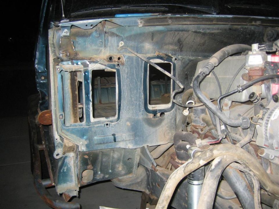 Teal vehicle firewall with two large rectangular cutouts, exposed engine components, and rusty bumper visible