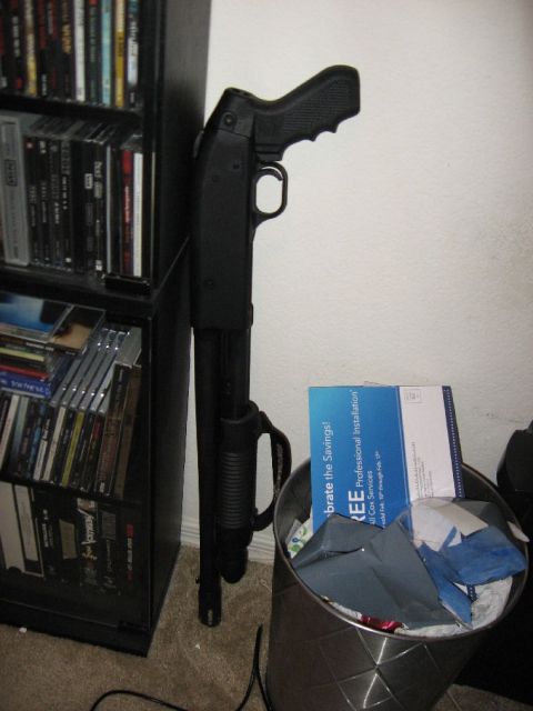 1978 Ford Bronco - A black shotgun is propped against a bookshelf, with a trash can filled with papers nearby.