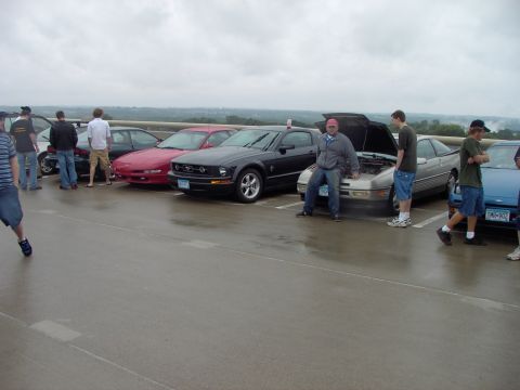 1989 Ford Probe - Several cars are parked in a lot, with people standing around them. One car has its hood open.