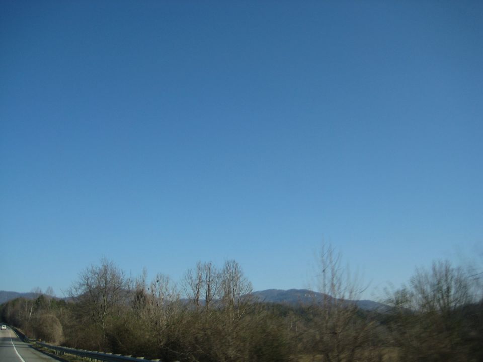 Roadside view of trees and mountains under a clear blue sky. Watermark reads SUPERMOTORS