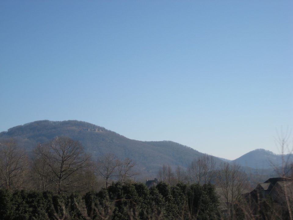 Mountain landscape with trees and houses under a clear blue sky. Watermark reads SUPERMOTORS