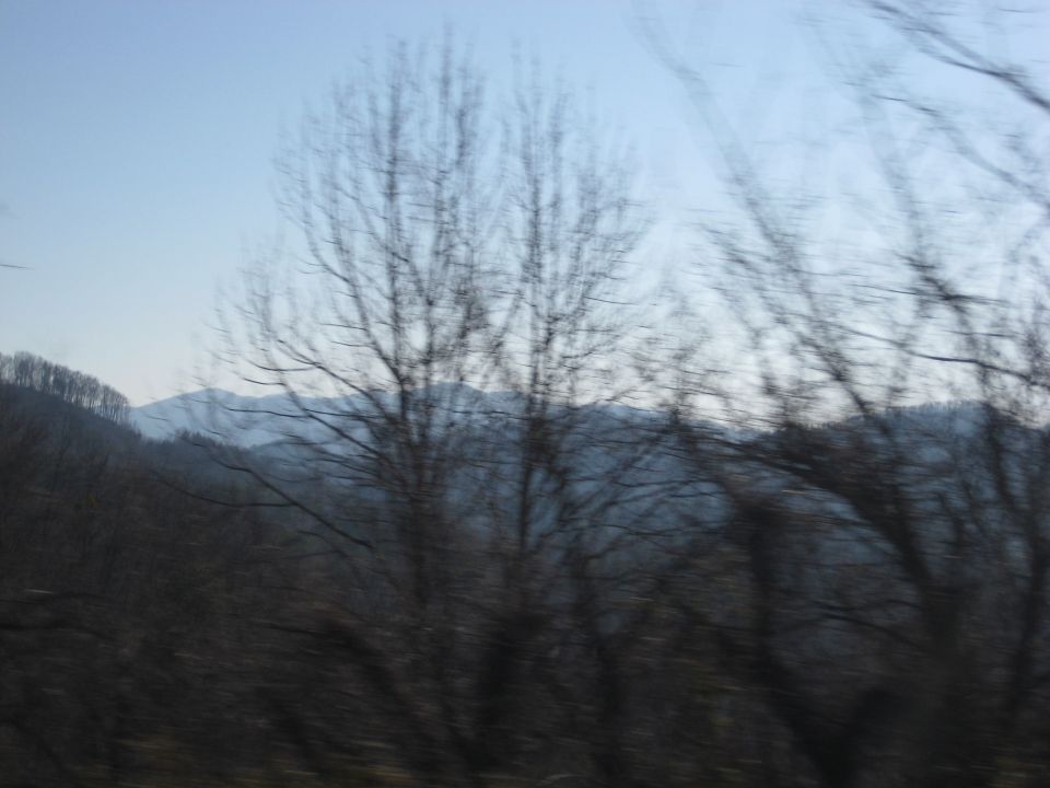 View from a moving vehicle shows bare trees and distant mountains under a hazy sky. Supermotors logo in the corner