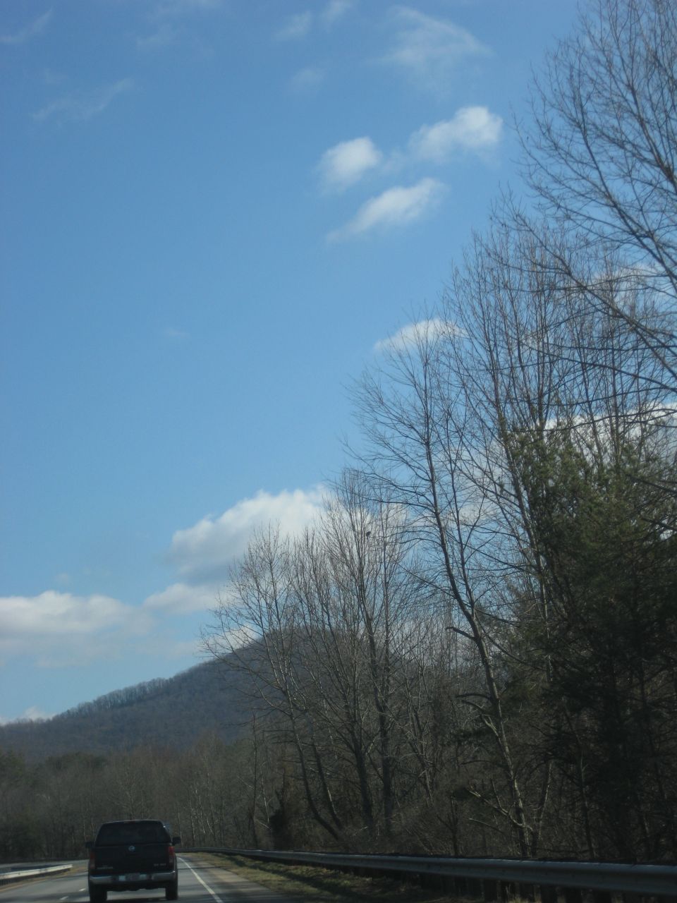 Truck driving on a highway with a wooded hillside in the background under a blue sky
