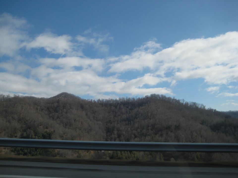Mountain landscape with bare trees under a blue sky with scattered clouds. A metal guardrail is in the foreground