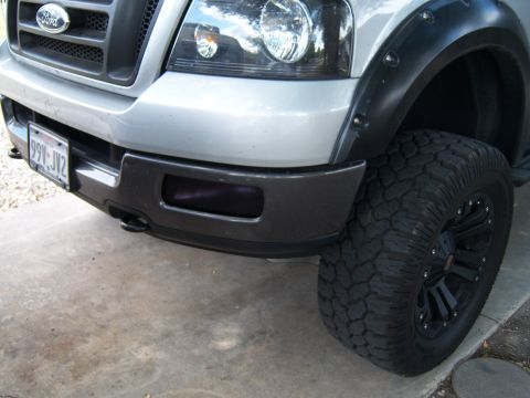 1979 Ford Bronco - Close-up of a silver Ford F-150 truck's front end, showing the grille, headlight, and a large off-road tire.