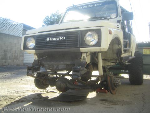 1987 Suzuki Samurai - Low front view of a white Suzuki Samurai up on stands mid-suspension work, with the front axle housing, leaf springs and brake drum backing…