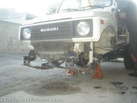 1987 Suzuki Samurai - A white 1987 Suzuki Samurai is shown with its front end raised on jack stands, revealing modifications to the axle.