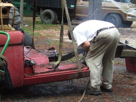 1967 Ford Bronco - A man works on a heavily rusted red vehicle chassis being lifted by straps, with other vehicles in the background.