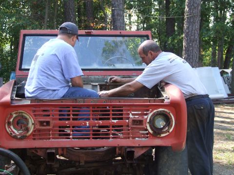 1967 Ford Bronco - Two men work on the engine bay of a red 1967 Ford Bronco, which is missing its grille and headlights.
