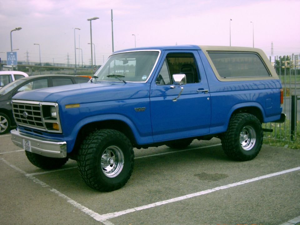 Blue 1982 Ford Bronco with tan roof and chrome wheels, parked in a lot.
