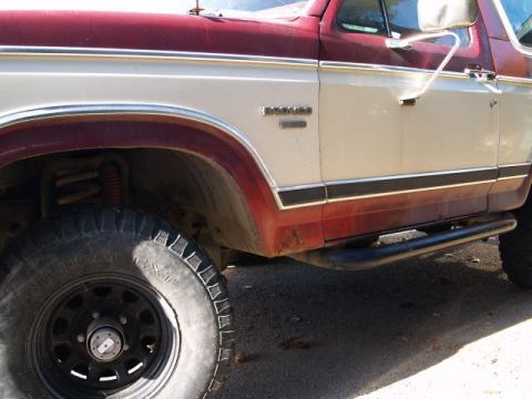 1996 Ford Bronco - A close-up side view of a two-tone red and white Ford Bronco shows the front fender, door, and running board. The vehicle has aftermarket b…