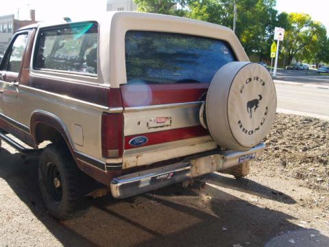 1996 Ford Bronco - The rear passenger side of a tan and maroon Ford Bronco is shown parked on a gravel shoulder next to a road. A spare tire cover with "Ford…