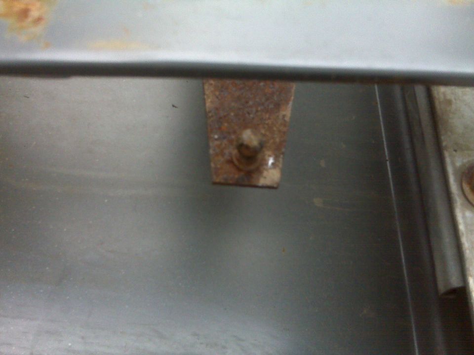 Rusty metal bracket with corroded bolt protruding, attached under a gray sheet-metal edge