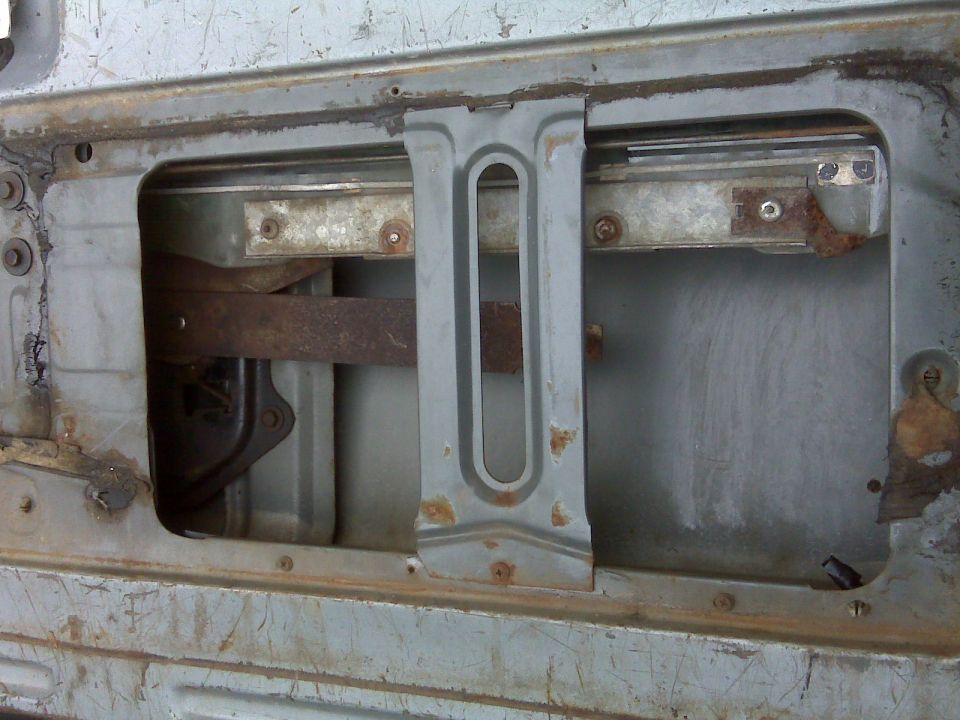 Inner door panel opening exposing window regulator mechanism and rails on a bare gray door with rust