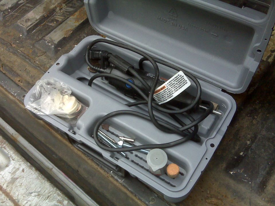 Open gray plastic case with rotary tool, coiled cord, flex shaft, and accessories sitting in a rusty truck bed
