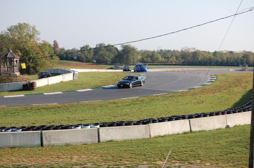 Dark BMW sedan on a race track with other cars, grass, and tire barriers.