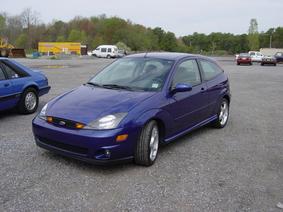 Purple Ford Focus hatchback parked in a gravel lot with trees behind
