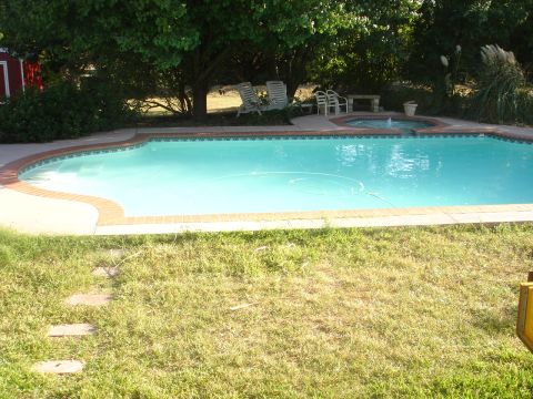 1989 Ford Bronco - A swimming pool with a brick border is surrounded by grass and trees. There is a small hot tub with a fountain in the background.