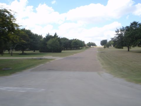 1989 Ford Bronco - A paved road curves through a park-like setting with manicured lawns and scattered trees under a partly cloudy sky.
