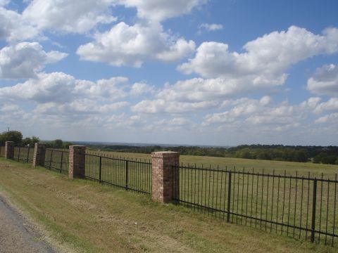 1989 Ford Bronco - A black metal fence with brick posts stands in a grassy field under a partly cloudy sky. The fence runs along a dirt road in the foreground…