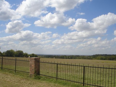 1989 Ford Bronco - A wide shot of a rural landscape under a cloudy blue sky. A black metal fence runs across the foreground, with a brick pillar supporting it…