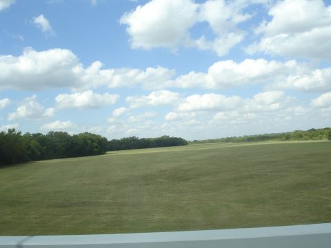 1989 Ford Bronco - A wide shot of a vast, grassy field under a partly cloudy blue sky. A dense line of trees borders the left side of the field, stretching in…