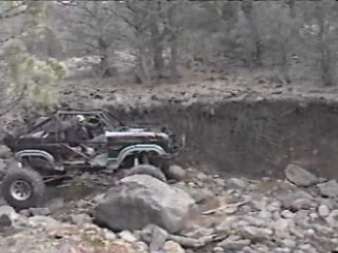 1984 Ford Bronco - A modified black Ford Bronco navigates a rocky, off-road terrain with a steep embankment to its right. The vehicle features a roll cage and…