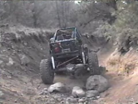 1984 Ford Bronco - A heavily modified off-road vehicle, likely a Jeep or similar, navigates a narrow, rocky trail with steep dirt embankments.