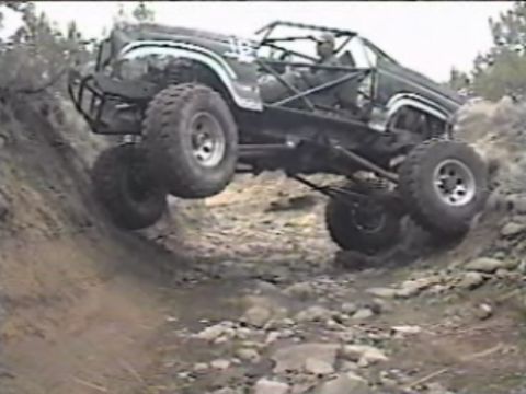 1984 Ford Bronco - A heavily modified dark-colored off-road vehicle, possibly a Ford Bronco, navigates a steep, rocky incline with its front wheels lifted hig…