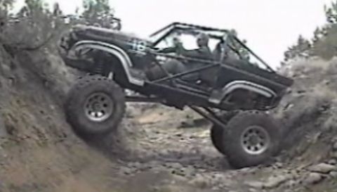 1984 Ford Bronco - A modified black Ford Bronco is shown at an extreme angle, climbing a steep, rocky incline off-road. The vehicle has a roll cage and large…