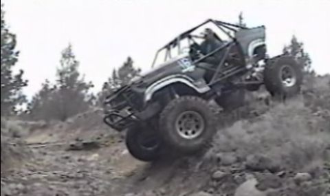 1984 Ford Bronco - A heavily modified off-road Ford Bronco is captured at a steep angle as it climbs a rocky incline, kicking up dust. The vehicle features a…
