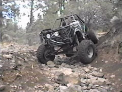 1984 Ford Bronco - A custom-built off-road vehicle, appearing to be a heavily modified 1984 Ford Bronco, navigates a steep, rocky incline in a wooded area. Th…