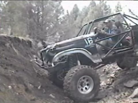 1984 Ford Bronco - A modified dark-colored Ford Bronco is navigating a steep, rocky incline with a driver and passenger visible inside. The vehicle has a roll…