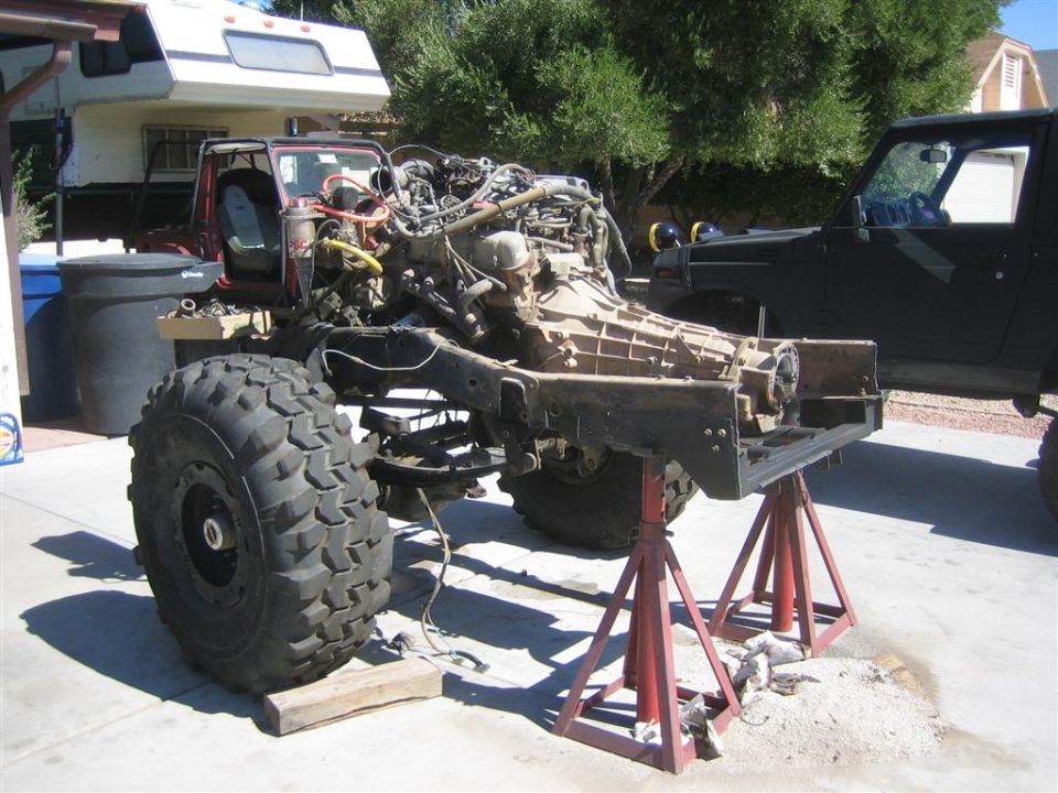 Vehicle chassis on jack stands, engine removed. Another vehicle is parked nearby