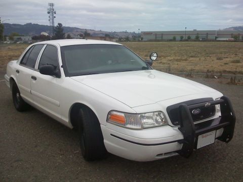 2000 Ford Crown Victoria - A white 2000 Ford Crown Victoria police car is parked in a lot with a large building in the background.