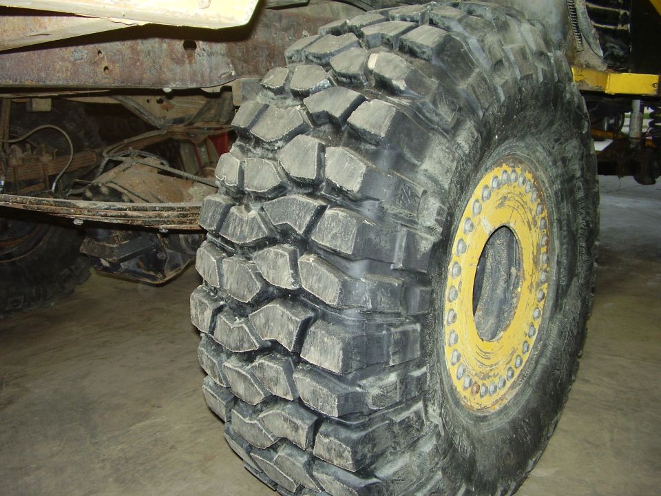 Off-road tire with aggressive tread and yellow beadlock wheel, showing leaf spring suspension and rusty frame