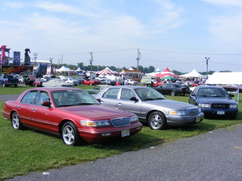 1997 Mercury Grand Marquis - A red 1997 Mercury Grand Marquis is parked on grass at an outdoor event, with other vehicles and tents in the background.