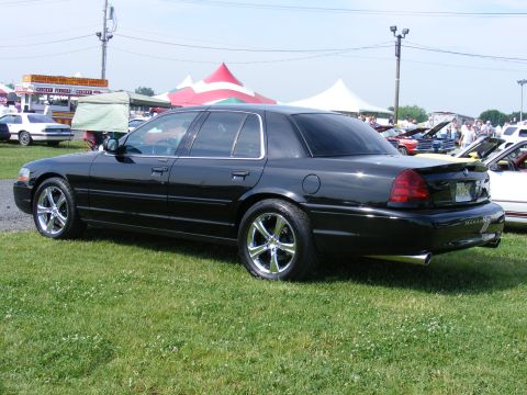 1997 Mercury Grand Marquis - A black sedan with aftermarket chrome wheels is displayed on a grassy field with tents and other vehicles in the background.