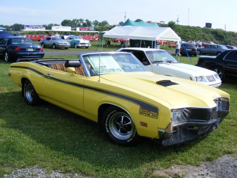 1997 Mercury Grand Marquis - Yellow 1970 Cyclone Spoiler convertible with black stripes and plaid interior at an outdoor car show.