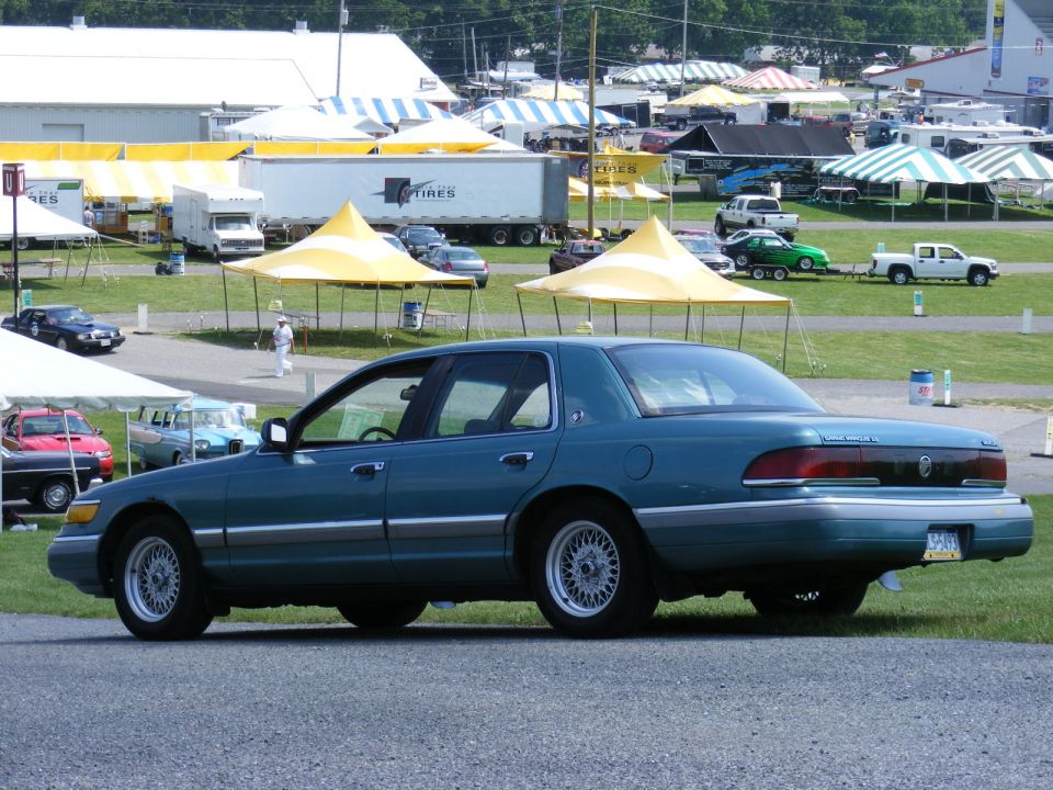 Teal sedan parked on asphalt, side view. Chrome trim along the body and silver wire wheels. Some tents and other vehicles in the background