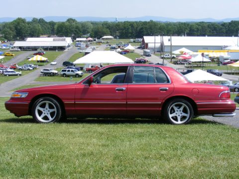 1997 Mercury Grand Marquis - A red 1997 Mercury Grand Marquis sedan is parked on a grassy area at an outdoor event with other vehicles and tents in the background.