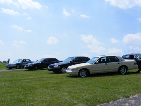 1997 Mercury Grand Marquis - A line of sedans, likely Ford Crown Victorias or Mercury Grand Marquis, are parked on a grassy field under a cloudy sky.
