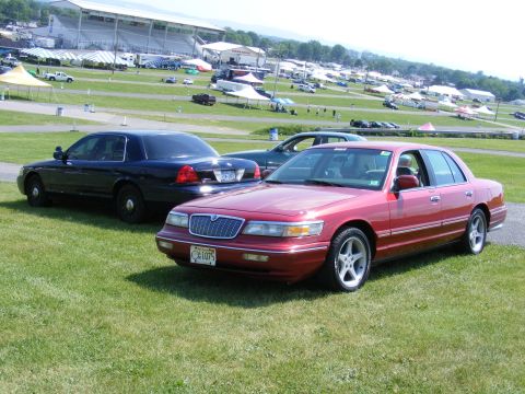 1997 Mercury Grand Marquis - Two sedans are parked on a grassy field with a stadium and tents in the background.