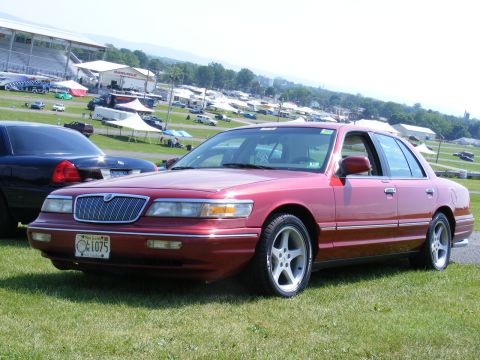 1997 Mercury Grand Marquis - A red sedan is parked on a grassy field with tents and bleachers in the background.